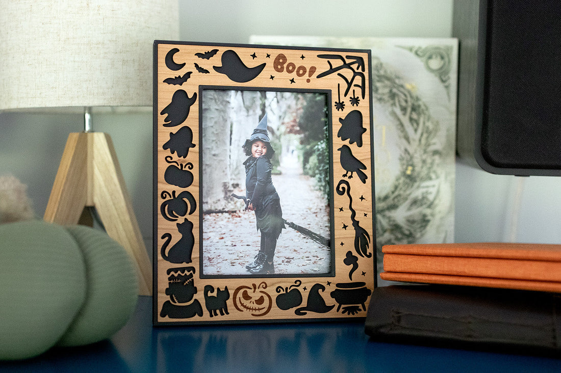 A laser-cut halloween picture frame for halloween keepsake and fall decor. The frame has a wood inlay and is sitting on a dresser next to a decorative pumpkin and photo albums.