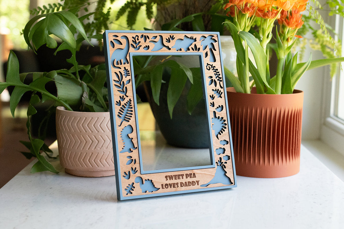 A handmade laser-cut dinosaur picture frame in slate blue and cherry wood, personalized with an engraved message, styled on a windowsill among plants — a kids' keepsake frame from BellaForm Creative's kids collection.