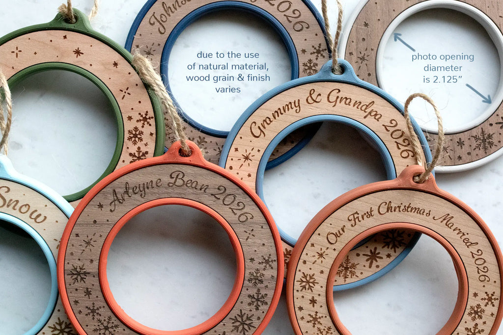 Personalized Christmas ornament frames without photos, showing laser-engraved names and snowflakes on maple, cherry, and walnut inlays in multiple housing colors. Caption text reads "photo opening diameter is 2.125 inches" and notes that wood grain and finish vary because the wood is natural.