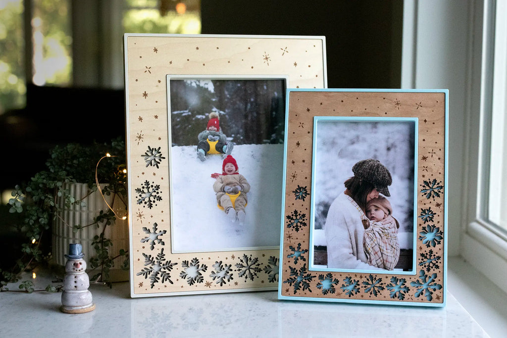 5x7 winter snowflake picture frame and 4x6 snowflake frame, sitting side-by-side, surrounded by Christmas lights and holiday decor, including a ceramic snowman. These keepsake winter frames are available in various sizes, colors, and orientations.