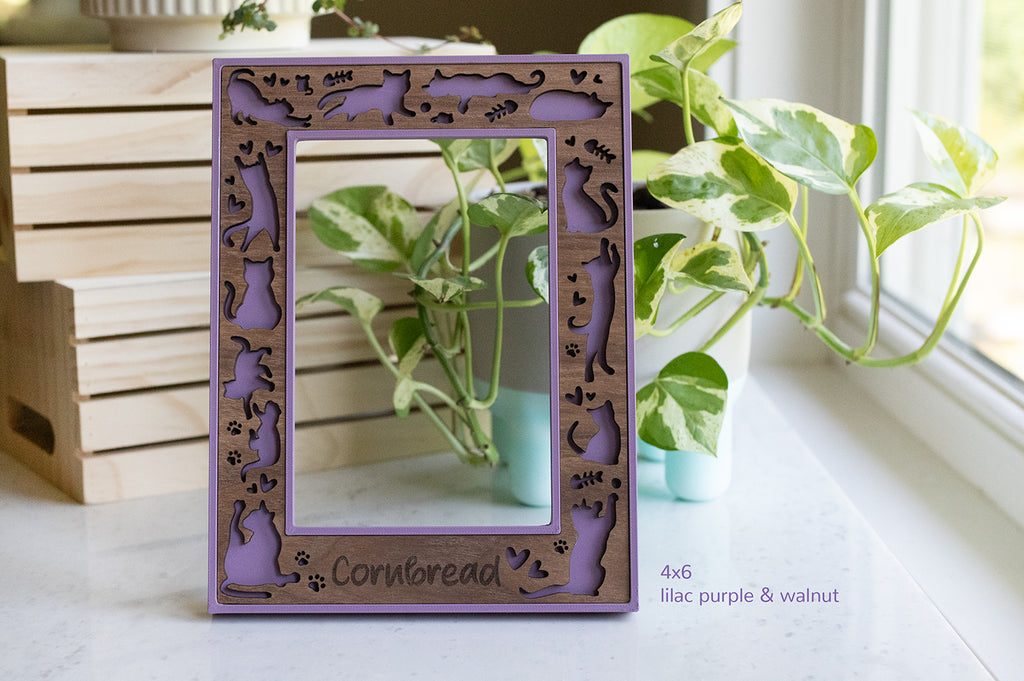 A 4x6 cat frame with laser cut design featuring cats, fish bones, mice, and little hearts. The frame is lilac purple with a walnut wooden inlay. The frame can be personalized with your cat's name or a custom message. There is greenery in the background.