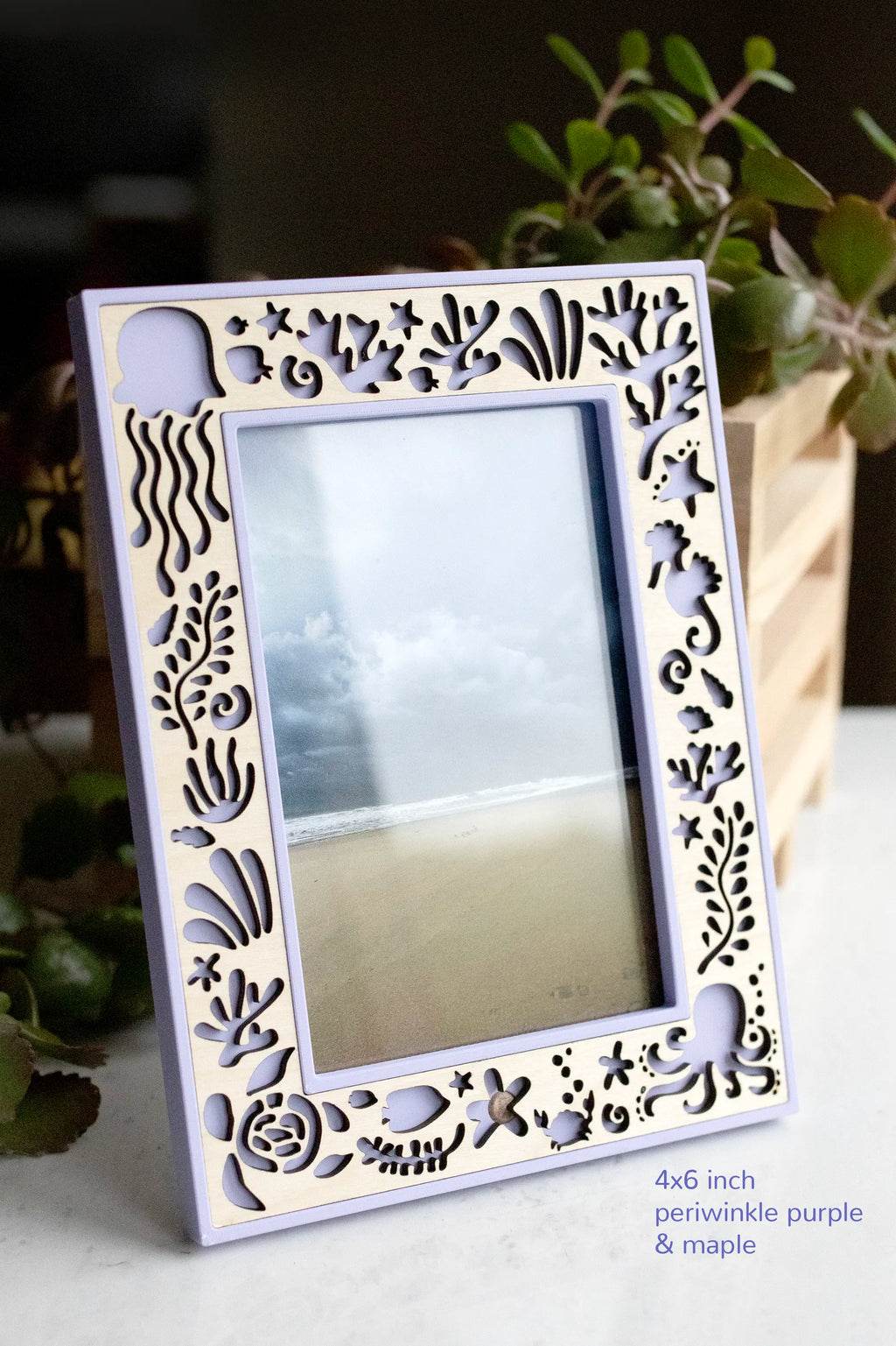 A purple and maple wood custom picture frame featuring laser cut sea creatures for an Under the Sea decor theme. Size is a 4x6 frame in vertical orientation, and it is displaying a photograph of the ocean. The frame is sitting on a white marble surface with greenery in the background.