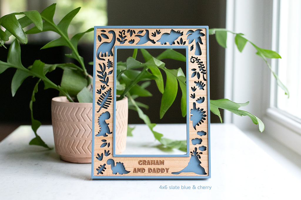 A custom 4x6 picture frame featuring a cute dinosaur theme. The frame border is slate blue with a wooden inlay made from cherry wood. Personalized with the message "Graham and Daddy". The frame is on a white surface with greenery in the background.