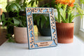 A custom engraved photo frame featuring a cute dinosaur theme. The frame border is blue with a wooden inlay made from cherry wood. Personalized with the message "Sweet Pea Loves Daddy". The frame is on a white surface with greenery in the background.