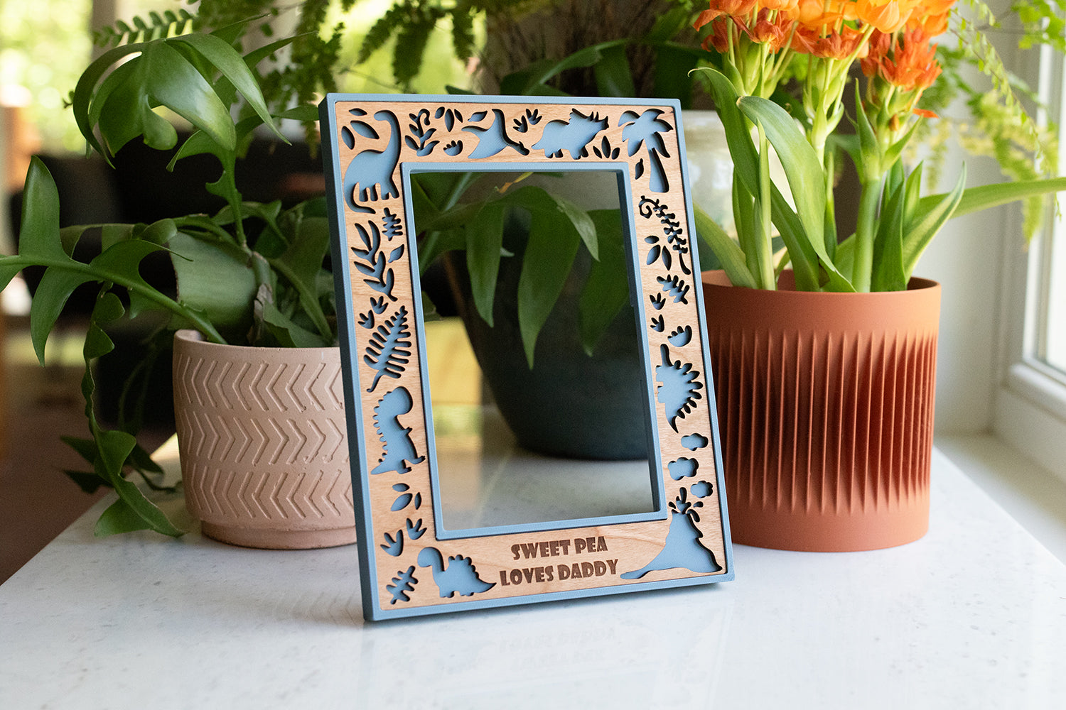 A custom engraved photo frame featuring a cute dinosaur theme. The frame border is blue with a wooden inlay made from cherry wood. Personalized with the message "Sweet Pea Loves Daddy". The frame is on a white surface with greenery in the background.