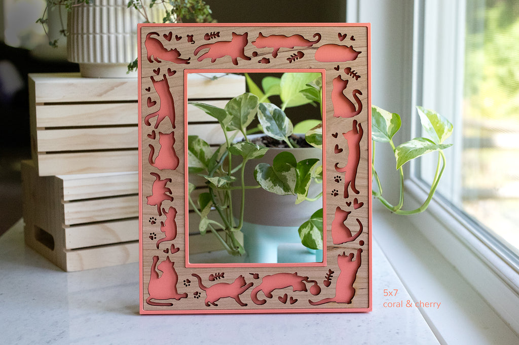 A whimsical cat picture frame with laser cut cat designs. The frame is coral pink with a cherry wood insert. The cats are playing, sleeping, loafing, and running around. There are little hearts, mice, and fish bones around the cats. The frame pictured is a 5x7 cat frame in vertical orientation.