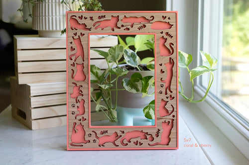 A whimsical cat picture frame with laser cut cat designs. The frame is coral pink with a cherry wood insert. The cats are playing, sleeping, loafing, and running around. There are little hearts, mice, and fish bones around the cats. The frame pictured is a 5x7 cat frame in vertical orientation.