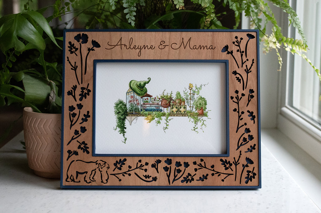 A personalized engraved photo frame featuring a baby bear hugging a mama bear, surrounded by woodland flowers. The frame has a navy blue border and laser cut cherry wooden inlay. The frame is displaying whimsical garden scene art in horizontal orientation.