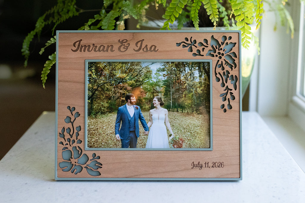 An elegant floral wood frame for a wedding keepsake. The frame is cherry wood and sage green with a delicate floral design around the border. The picture frame holds a 5x7 photograph, and it has a horizontal orientation. The frame displays a photograph of a couple at their wedding.