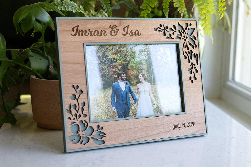 A delicate floral wood frame for a wedding keepsake. The frame is cherry wood and sage green with a delicate floral design around the border. The green frame is 3D printed, and the wooden decorative inlay is laser cut from real wood. The frame is sitting on a white marble surface.