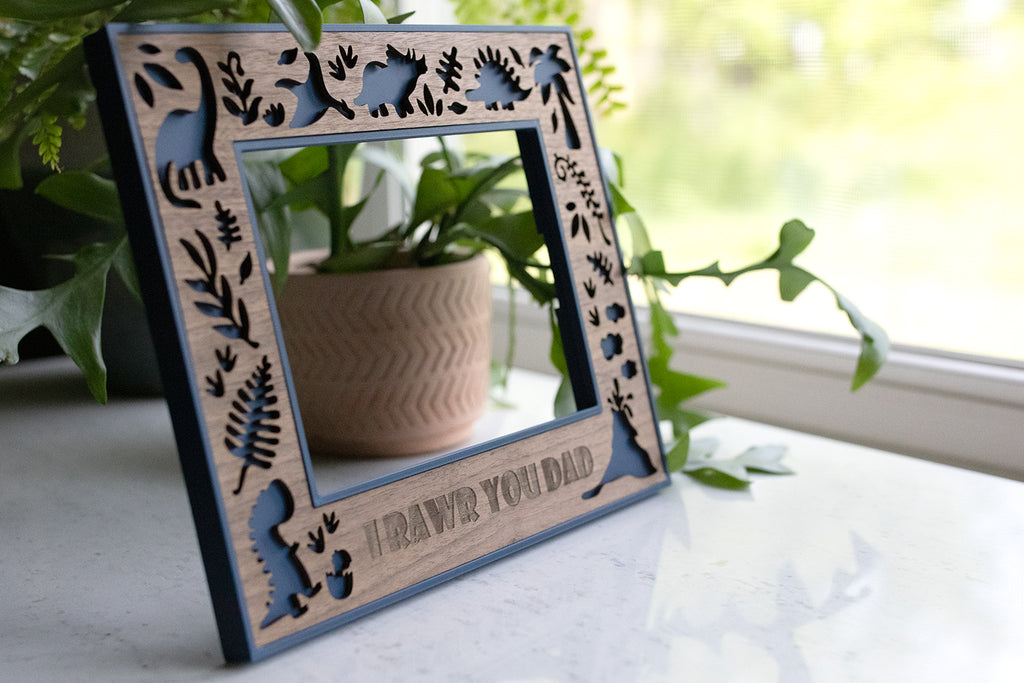 Wooden picture frame with a navy blue border and cute dinosaur cutouts. The frame is customized with the message "I RAWR YOU DAD" and is decorated with plant and dinosaur silhouettes. The frame is displayed near a window with plants.