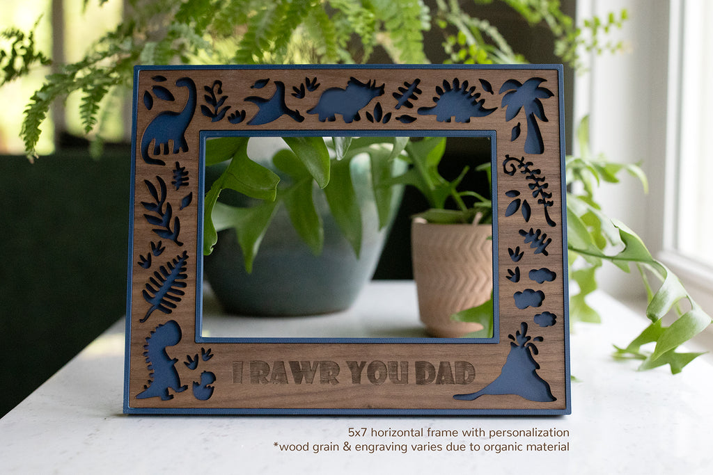 A wooden dinosaur picture frame with a blue border and dinosaur cutouts. The frame has the phrase "I RAWR YOU DAD" engraved at the bottom. The frame is 5x7 inches.