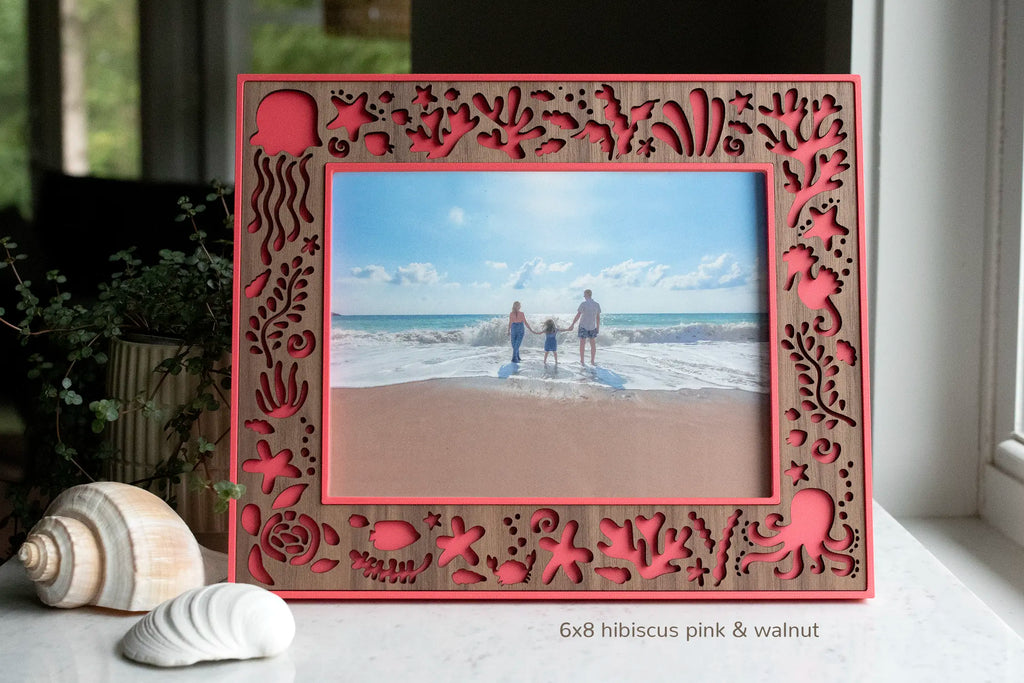 A large coastal picture frame that features laser-cut ocean creatures and under the sea elements. The frame holds a 6x8 photograph, has a "hibiscus pink" frame housing with an intricately cut walnut inlay. Designed to frame your family's favorite beach vacation photo for beach house decor or a summer keepsake. 