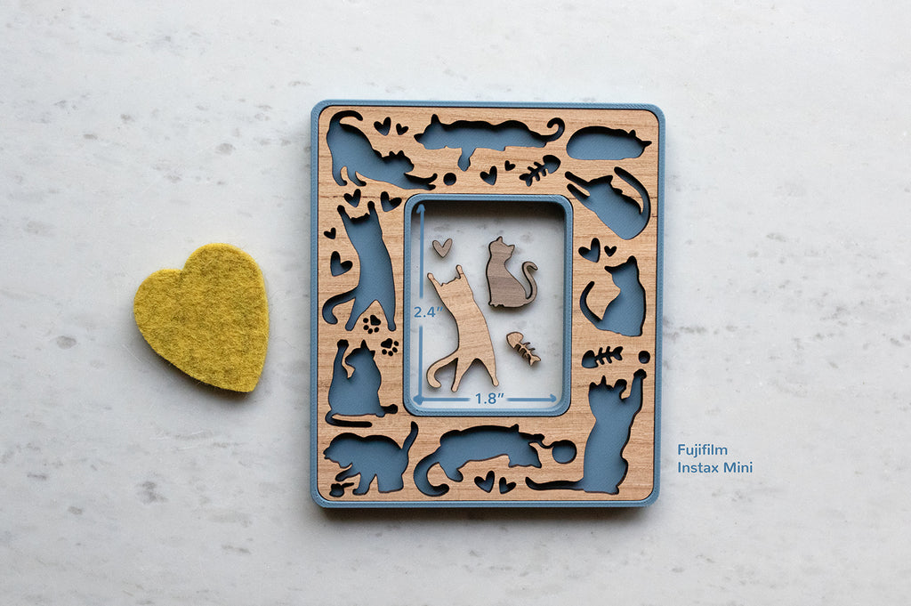 A blue instax mini cat frame with cute laser cut cats in a cherry wood inlay. The frame is lying on a white surface next to a yellow felt hearts. The frame features cats playing, stretching, and sleeping along with little mice, hearts, paw prints, and fish bones. The photo opening in the frame is 2.4" x 1.8".