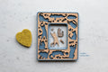 A blue instax mini cat frame with cute laser cut cats in a cherry wood inlay. The frame is lying on a white surface next to a yellow felt hearts. The frame features cats playing, stretching, and sleeping along with little mice, hearts, paw prints, and fish bones. The photo opening in the frame is 2.4" x 1.8".