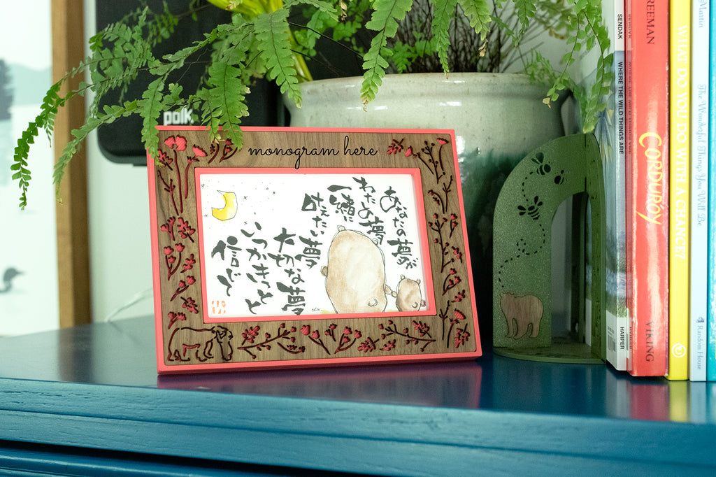 Hibiscus pink and personalized laser cut walnut inlay photo frame displaying a piece of Japanese art with a mama bear holding hands with a baby bear. In the background is a potted green fern and a 3D printed bear themed bookend holding up a few children's' books. The items are sitting on a turquoise blue wooden cabinet.