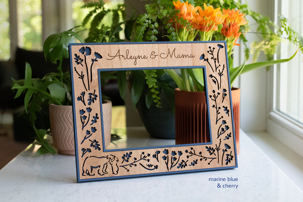 An engraved photo frame featuring a baby bear hugging a mama bear, surrounded by woodland flowers. The frame has a navy blue border and laser cut cherry wooden inlay. Custom frames are available in 5x7, 4x6, Polaroid Instax Wide, and ultrasound frame sizes.