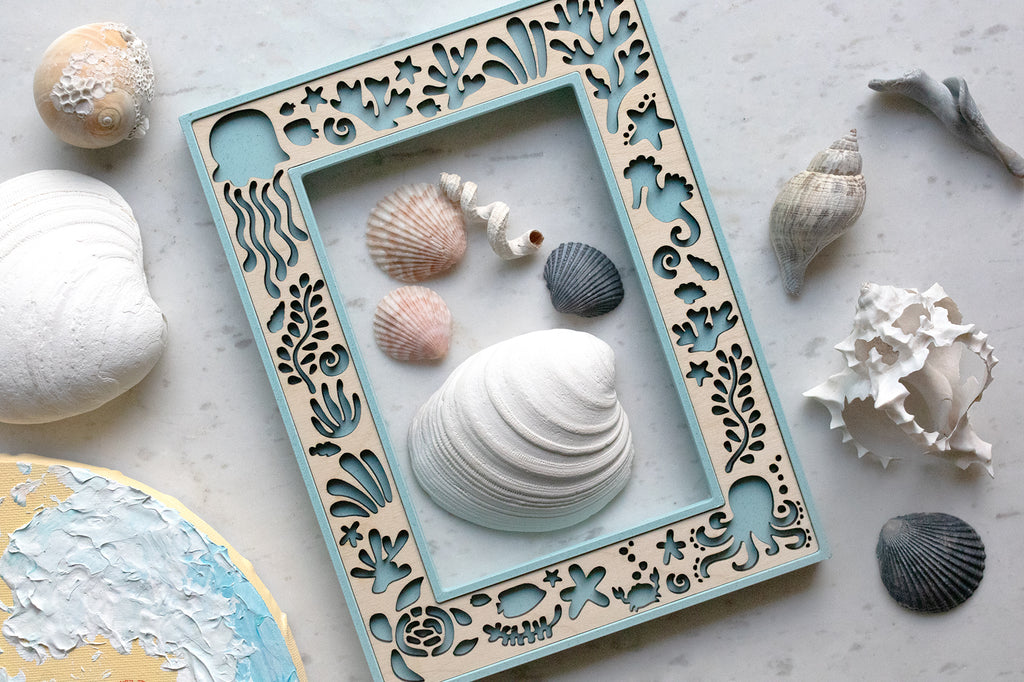Handmade keepsake ocean creatures picture frame featuring laser-cut marine motifs including jellyfish, seahorses, crabs, and seaweed. Several seashells of varying sizes, shapes, and colors are arranged around and inside the frame.