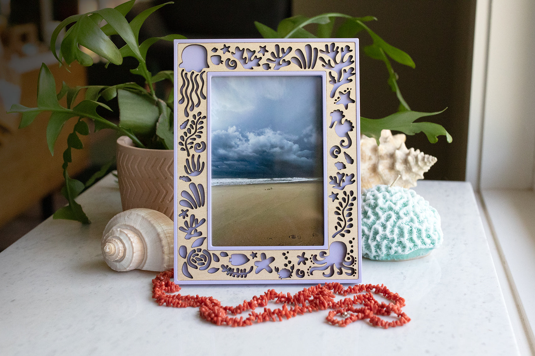 A light purple and wooden custom picture frame featuring laser cut sea creatures for an Under the Sea decor theme. Size is a 4x6 frame in vertical orientation, and it is displaying a photograph of the ocean. The frame is sitting on a white marble surface with a coral necklace, seashells, and greenery in the background.
