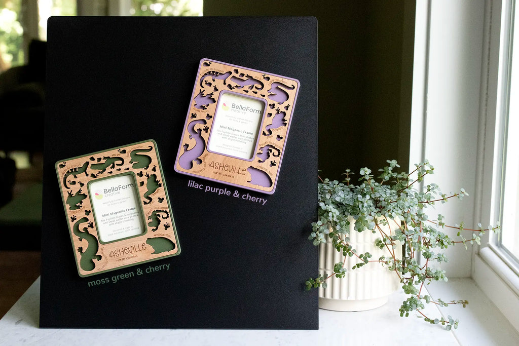 Two magnetic Appalachian amphibians frames in moss green and lilac purple with cherry wood inlay, laser-cut salamander, frog, and toad cutouts with engraved species names, and “Asheville, North Carolina” engraving, displayed on a black magnetic board.