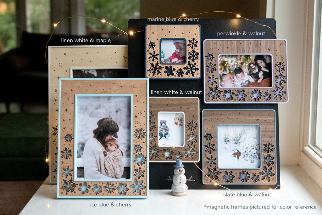 An assortment of snowflake holiday picture frames with various colors and wood types. Frames are laser cut with delicate snowflake designs and engraved falling snow. Magnetic frames are pictured for color reference. 4x6, 5x7, and 6x8 sizes available in the shop.