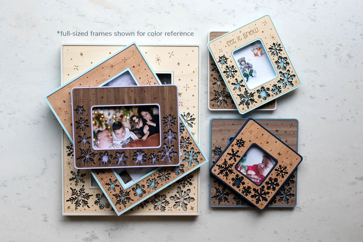 An assortment of snowflake picture frames in different colors and wood inlays, arranged on white marble and holding family photos — full-size frames shown here for color reference. Each is laser-cut with a lightweight, durable 3D-printed housing and can be customized by wood type and color. Available in 4×6, 5×6, Instax Mini, Instax Wide, and Polaroid sizes.