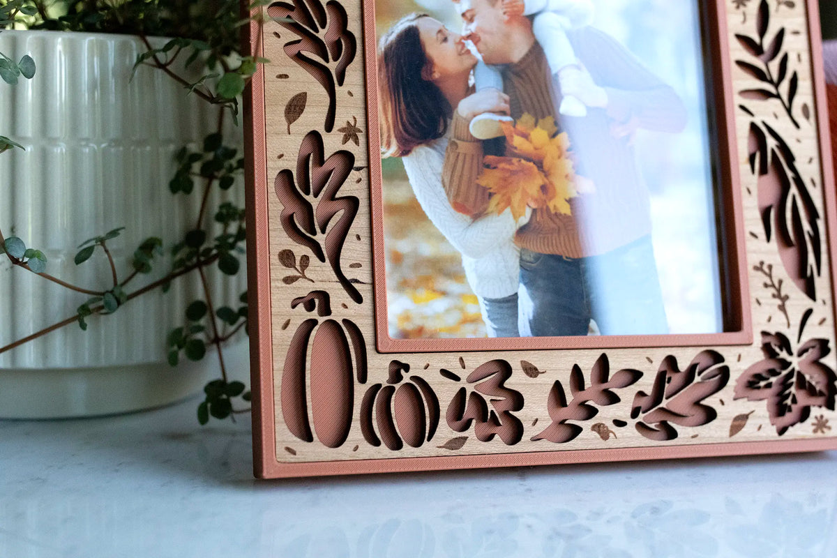 Detail of a terracotta fall picture frame with a laser-cut cherry wood inlay, featuring pumpkins, oak and maple leaves, acorns, and branches around a photo of a couple's fall photo. A personalized autumn keepsake and fall home decor piece, styled on a marble surface beside a white ribbed planter with trailing greenery.