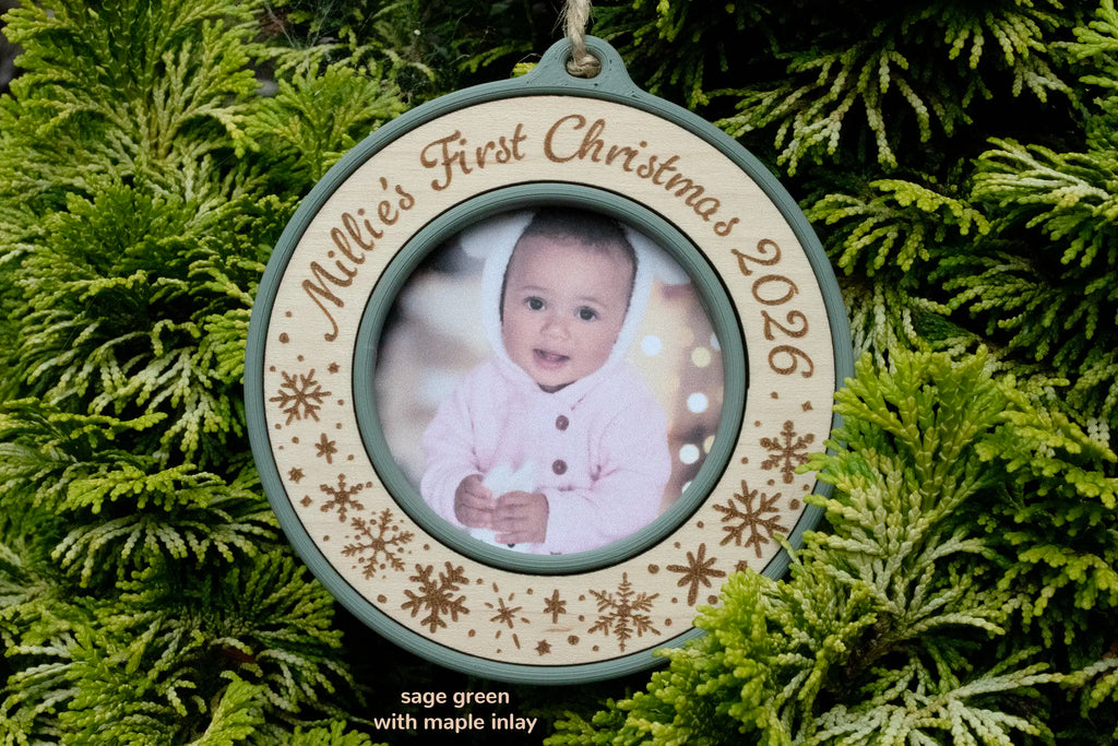 Baby's first Christmas photo ornament in sage green with a maple inlay, laser-engraved "Millie's First Christmas 2026" with snowflakes, holding a photo of a baby in a pink hooded outfit in front of the Christmas tree.