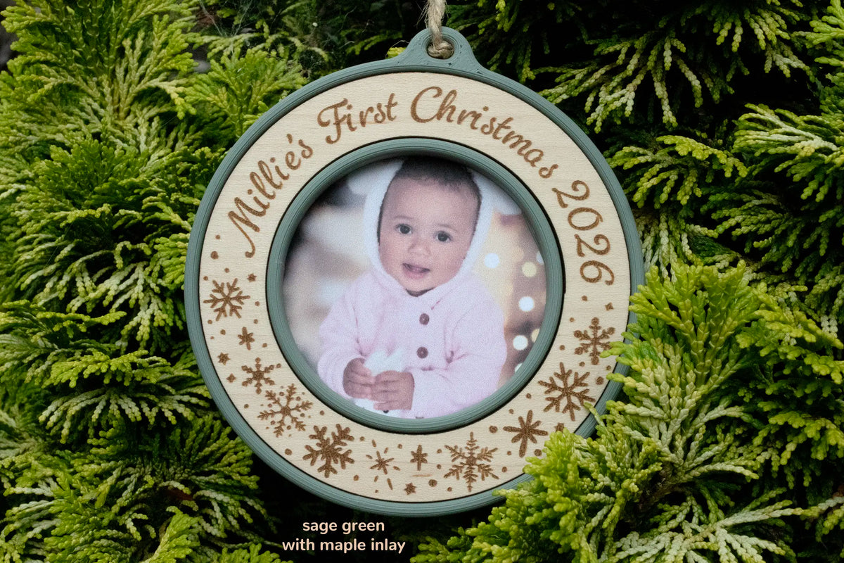 Baby's first Christmas photo ornament in sage green with a maple inlay, laser-engraved "Millie's First Christmas 2026" with snowflakes, holding a photo of a baby in a pink hooded outfit in front of the Christmas tree.