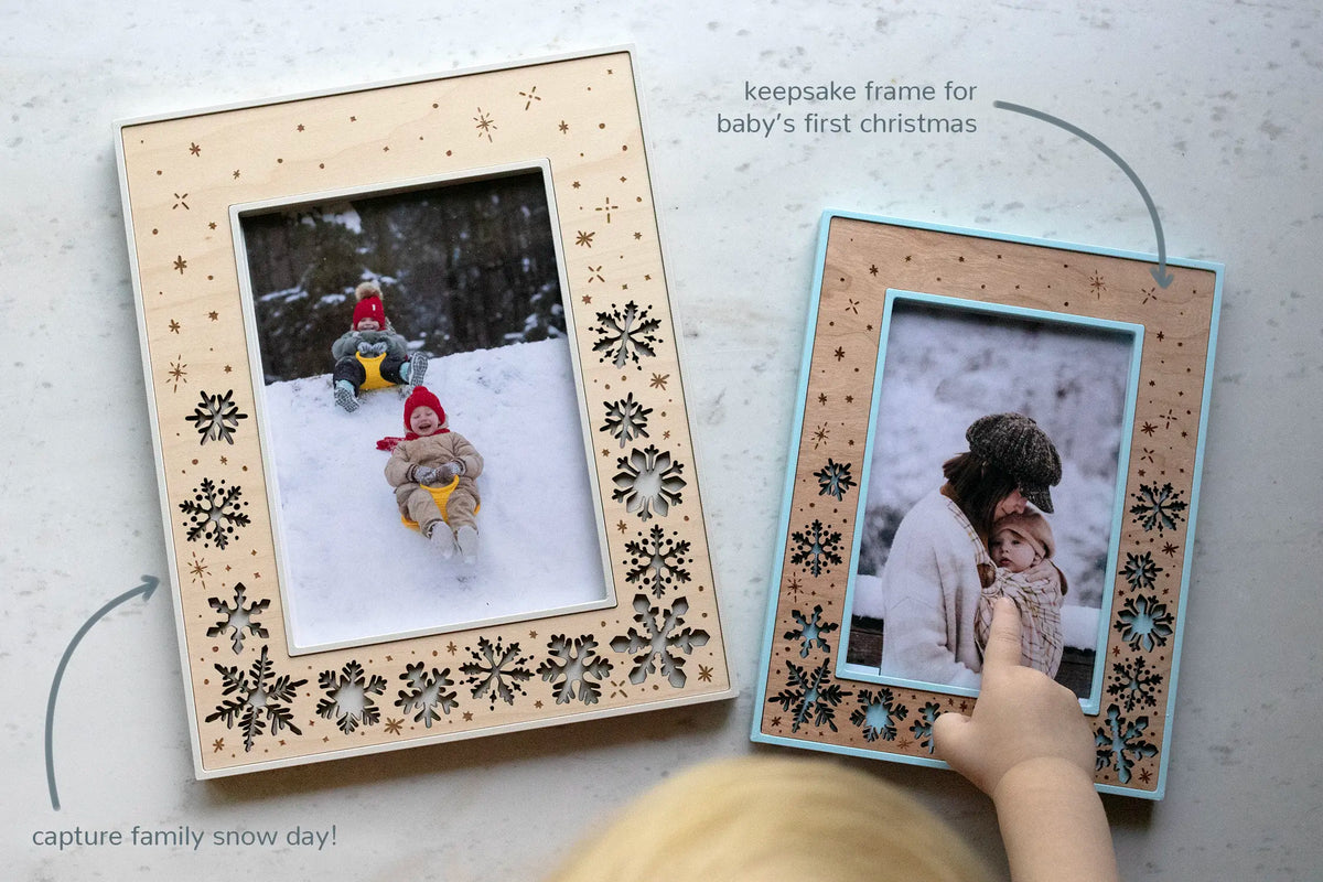Two Christmas holiday picture frames featuring laser cut snowflakes and bits of falling snow. Available in various shades of blue and linen white with three wood types, including walnut, maple, and cherry. The right frame shows a mom holding her baby in the snow with the caption "keepsake frame for baby's first Christmas". 