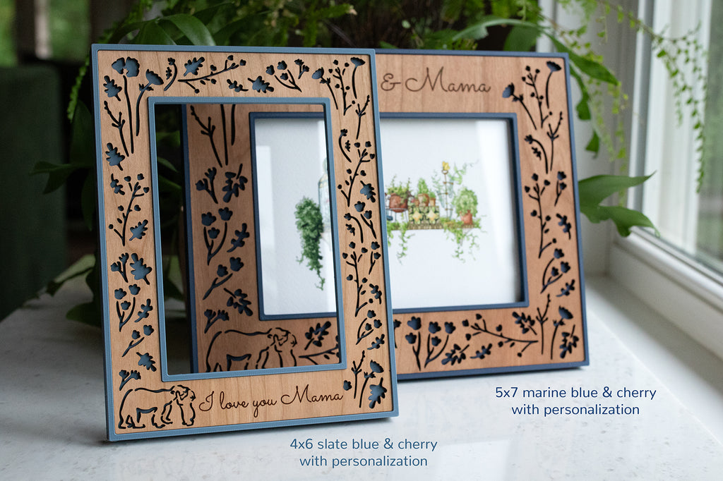 A navy blue and cherry wood 5x7 picture frame behind a slate blue and cherry 4x6 picture frame on a white marble surface. The laser cut frames feature whimsical woodland flowers with a baby bear hugging its mama bear. There is greenery in the background for a natural aesthetic.