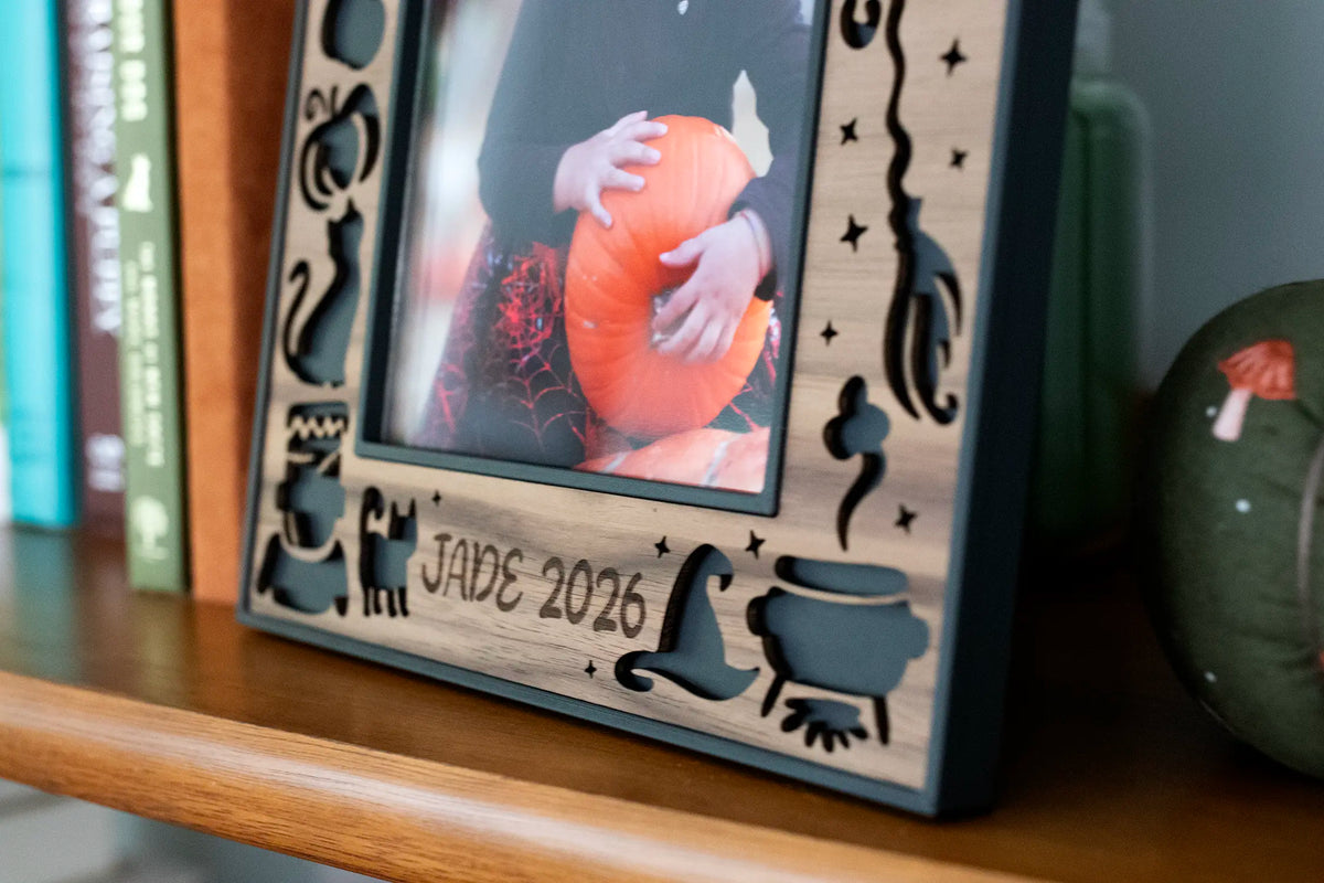 Close-up of a personalized Halloween picture frame with "Jade 2026" laser-engraved in walnut, framed by laser-cut witch hat, cauldron, black cat, and stars