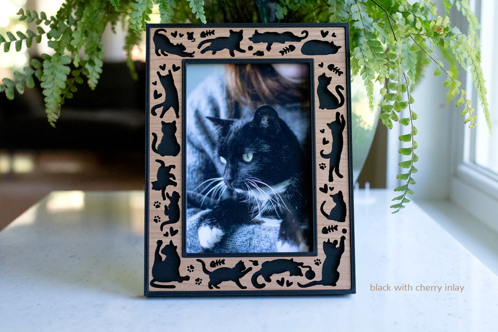 Black laser-cut 4x6 cat picture frame with a cherry wood inlay, bordered by cat silhouettes, paw prints, and hearts, displaying a photo of a black cat on a countertop.