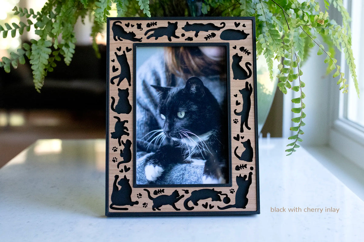 Black laser-cut 4x6 cat picture frame with a cherry wood inlay, bordered by cat silhouettes, paw prints, and hearts, displaying a photo of a black cat on a countertop.
