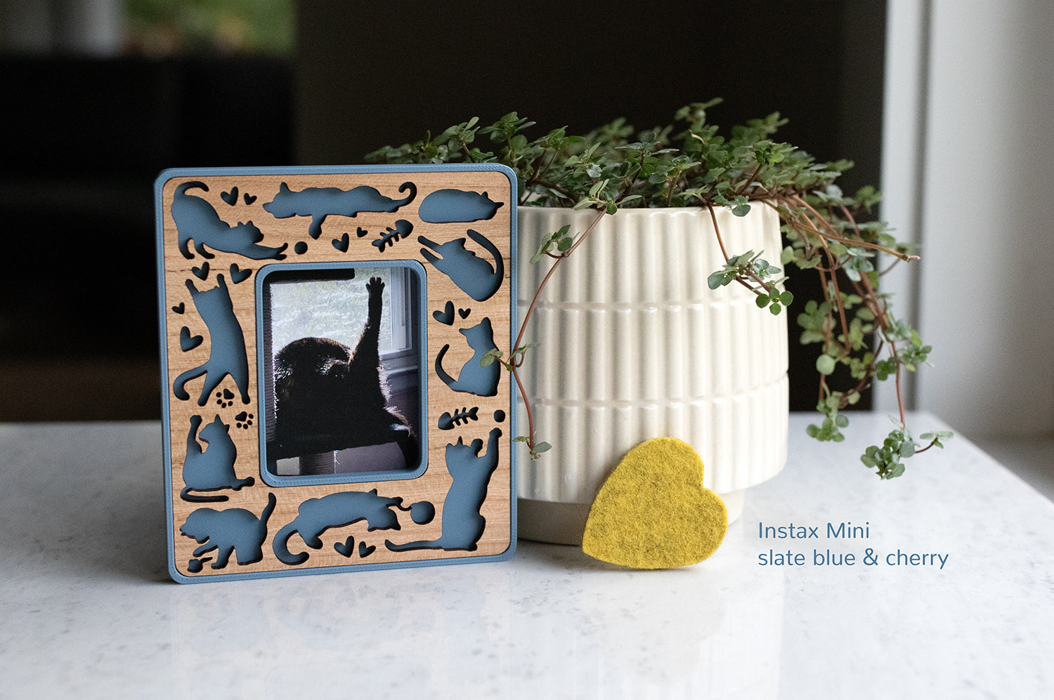 Instax Mini magnetic cat frame for cat lovers and cat memorial gifts. The frame is blue with a cherry wood laser cut inlay. It is propped up next to a white planter and a small yellow felt heart.