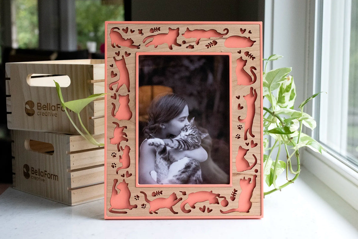 Coral cat picture frame with a laser-cut cherry wood border of cat silhouettes, paw prints, fish bones, and hearts, holding a black-and-white photo of a girl hugging her tabby cat, on a sunny windowsill beside a pothos plant.