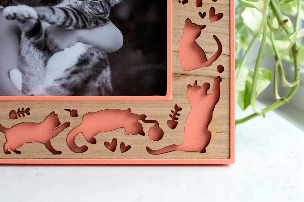 Close-up of the corner of a handcrafted cat lover picture frame in coral and cherry wood, showing laser-cut silhouettes of playful cats batting a ball of yarn, sitting and stretching, with fish bones and hearts cut into the wood border of this 3D-printed pet photo frame.
