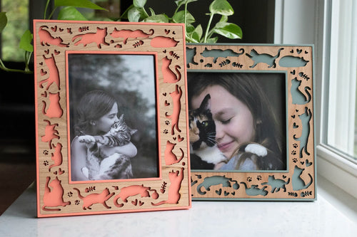 Two cat picture frames in coral and sage green, with laser-cut cherry wood borders of cat silhouettes, paw prints, and hearts. The vertical frame holds a black-and-white photo of a girl hugging her tabby cat, and the horizontal pet photo frame shows a girl cuddling a calico cat.