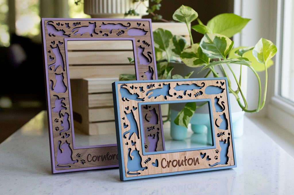 Two laser-engraved, personalized cat frames with the names "Cornbread" and "Crouton" engraved on the wood. Frames are purple and blue with walnut and cherry inserts.