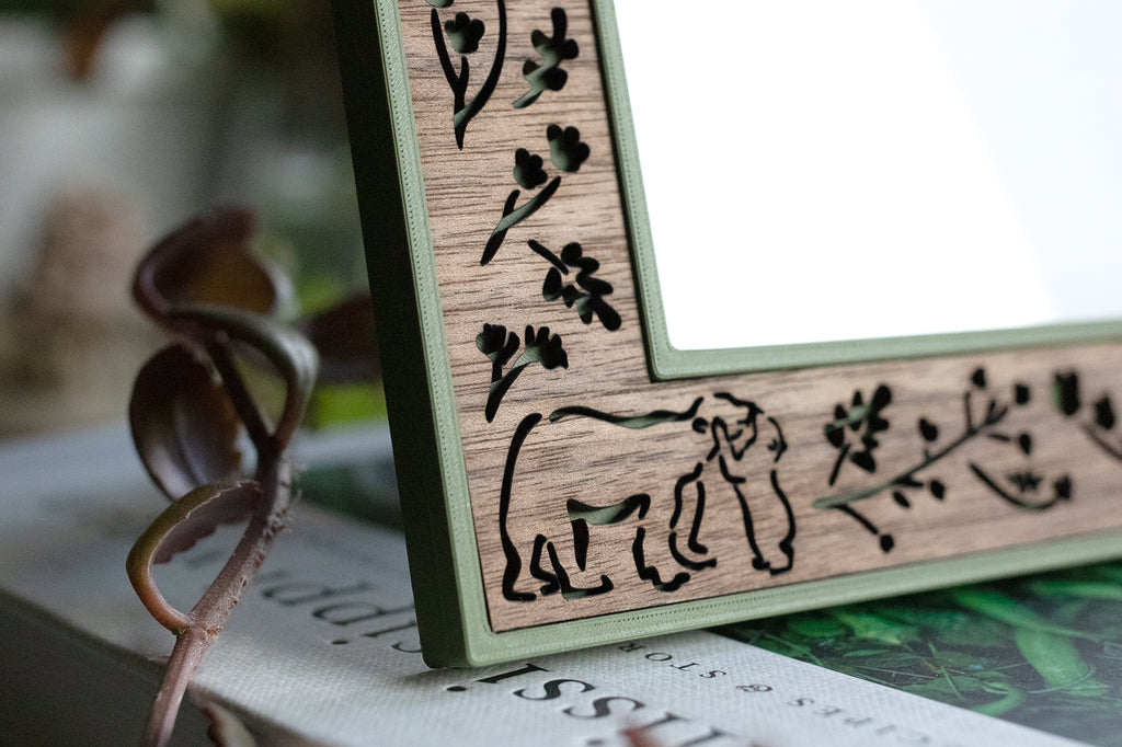 Close-up of the corner of an intricate walnut laser cut and green border picture frame. The laser cut details show a baby bear reaching up and giving mama bear a kiss. The bears are surrounded by laser cut flowers and leaves.
