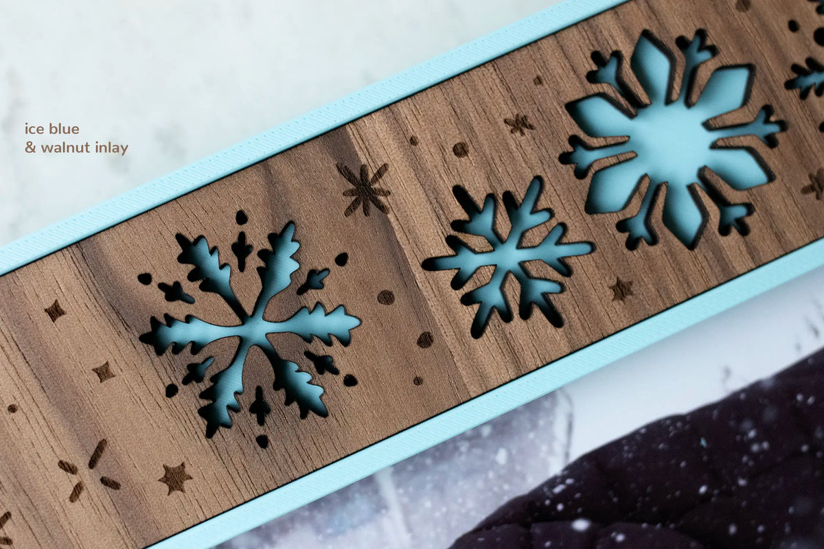 Close-up of the frame's walnut wood inlay with laser-cut snowflakes showing ice blue backing, plus engraved falling snow. Text reads "ice blue & walnut inlay."