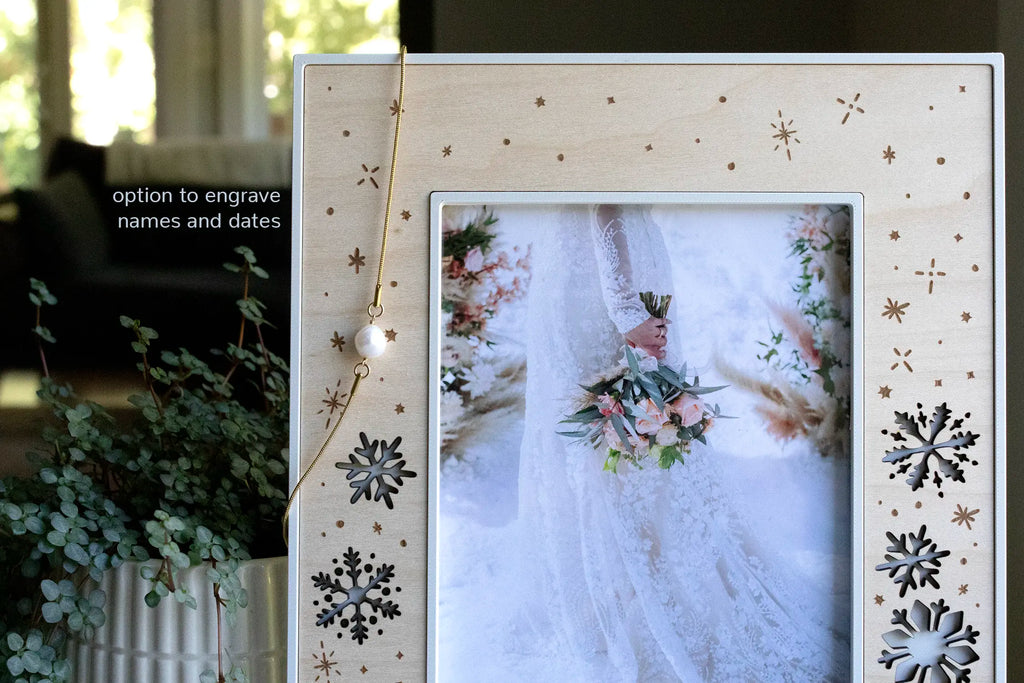 A close-up view of a laser-cut snowflake picture frame for a winter wedding keepsake. This wedding frame displays a photo of a bride holding a bouquet in the snow at her wedding. There is a gold necklace with a pearl hanging on the frame, and the words "option to engrave names and dates" are visible.