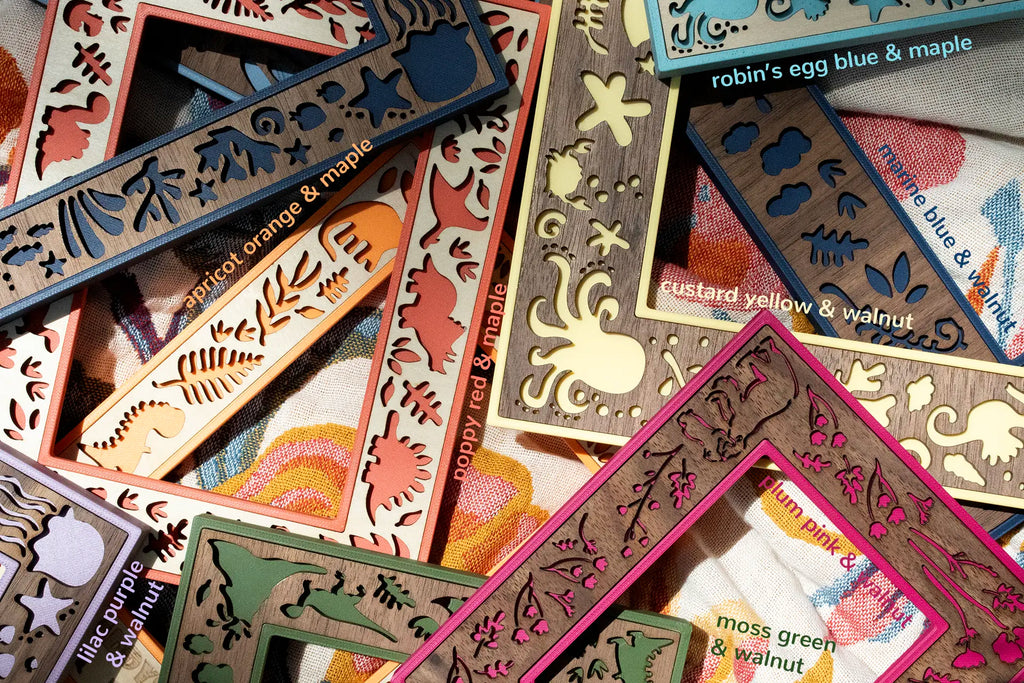 A collection of laser cut 3D printed and wooden frames in various colors, including apricot orange, poppy red, lilac purple, and moss green. Each frame features intricate cut-out designs of plants, animals, and sea creatures. The frames are labeled with color combinations such as "robin's egg blue & maple".