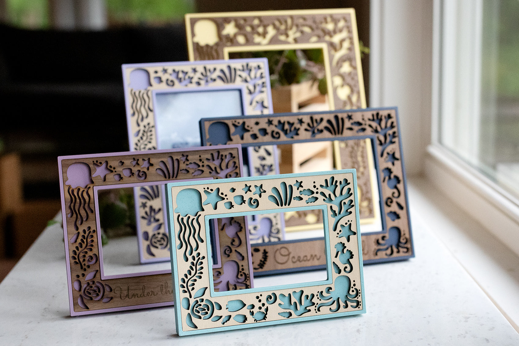 An assortment of custom wooden frames displaying ocean creatures and bright colors as well as walnut, birch, and cherry wooden inlays. The frames are photographed in natural sunlight and sitting on a white marble surface.