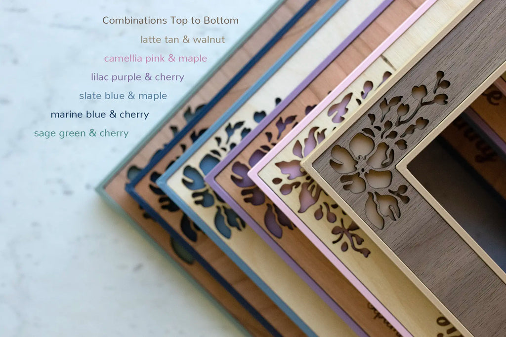 A top corner detail view of six floral wedding photo frames. On top is a tan and walnut frame, followed by pink and maple, lilac purple and cherry, slate blue and maple, marine blue and cherry, and sage green and cherry. The floral cut-outs are visible as the frames are stacked.