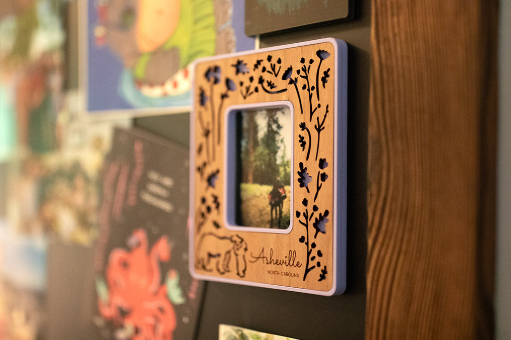 A close-up detail view of a laser engraved customizable Polaroid frame hanging on a magnetic chalkboard. The frame has a cherry wood inlay and is hanging next to a few greeting cards.