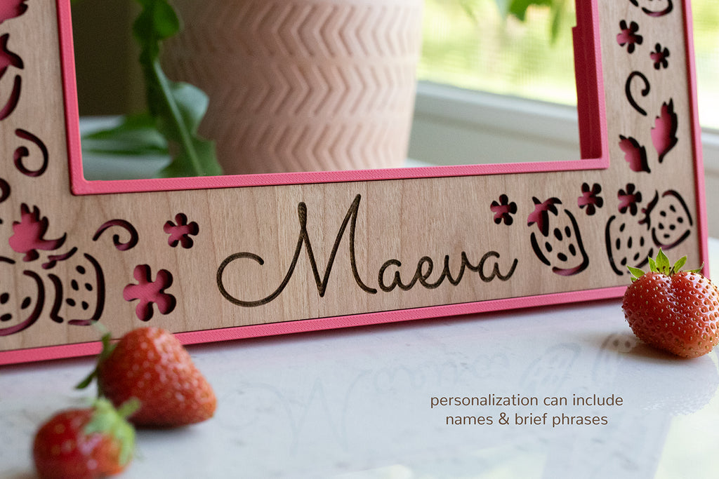 A detail view of the name "Maeva" laser engraved on a personalized picture frame. The frame features delicate strawberries and a bright pink frame with cherry wood inlay. It sits on a white surface with strawberries in the foreground.