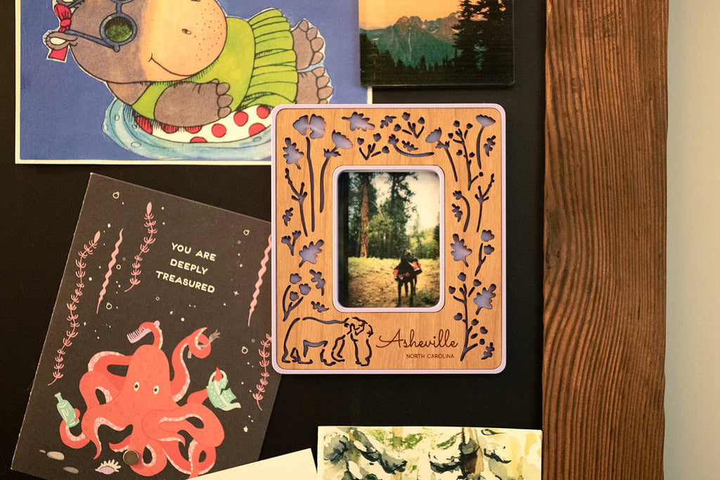 A close-up detail view of a laser engraved customizable Polaroid frame hanging on a magnetic chalkboard. The frame has a cherry wood inlay and is hanging next to a few greeting cards.