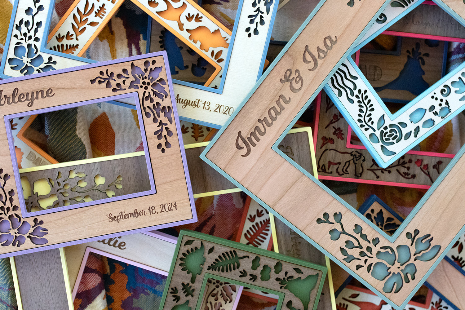 An assortment of colorful picture frames shown in various colors, wood styles, and sizes. The frames are pictured in natural sunlight. Personalized wood engravings with names and dates as well as laser cut flowers, sea creatures, and dinosaurs are visible.