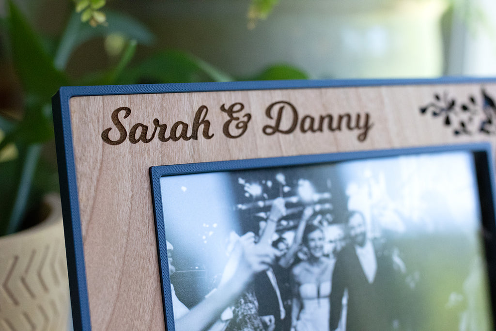 A close-up detail view of a 4x6 picture frame displaying "Sarah & Danny" engraved in the wood in a scripted font. The frame is navy blue and cherry wood with a floral design and is intended as a wedding gift.